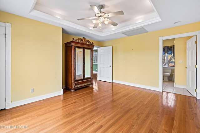 unfurnished bedroom featuring a tray ceiling, ceiling fan, ensuite bathroom, and light hardwood / wood-style floors