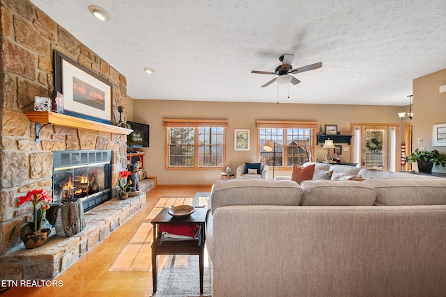 living room with ceiling fan with notable chandelier, a textured ceiling, light wood-type flooring, and a stone fireplace