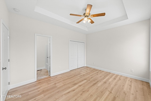 unfurnished bedroom featuring light hardwood / wood-style flooring, ceiling fan, connected bathroom, a tray ceiling, and a closet