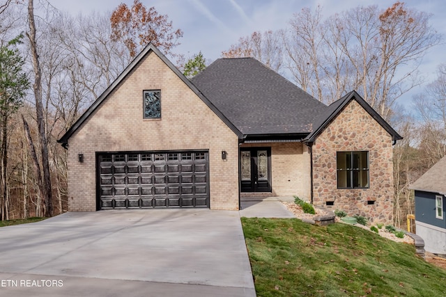 french country style house with driveway, stone siding, roof with shingles, a garage, and brick siding