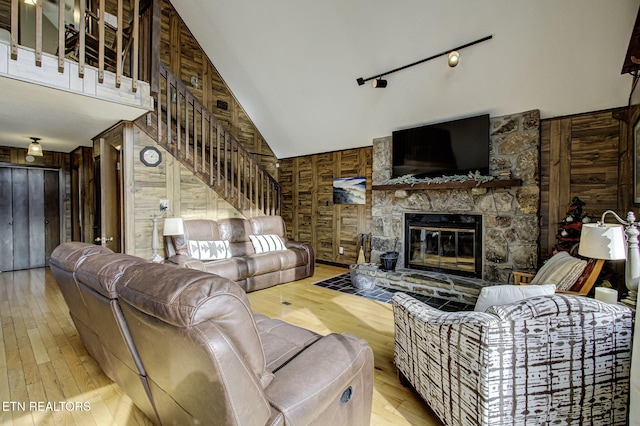 living room featuring rail lighting, a stone fireplace, high vaulted ceiling, light hardwood / wood-style floors, and wooden walls