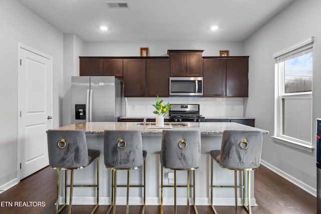 kitchen featuring stainless steel appliances, light stone counters, dark hardwood / wood-style flooring, an island with sink, and a kitchen bar