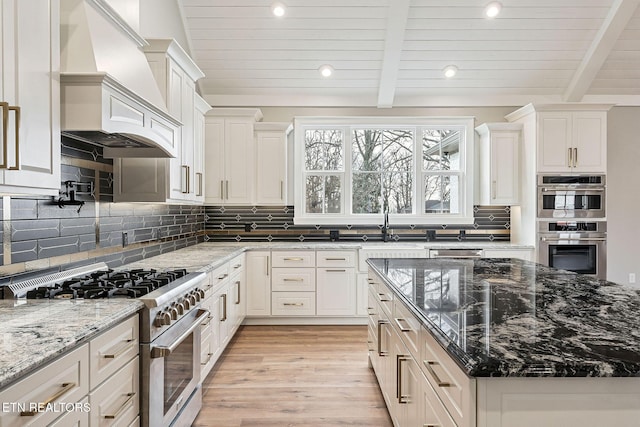 kitchen featuring tasteful backsplash, custom range hood, stainless steel appliances, dark stone countertops, and white cabinetry