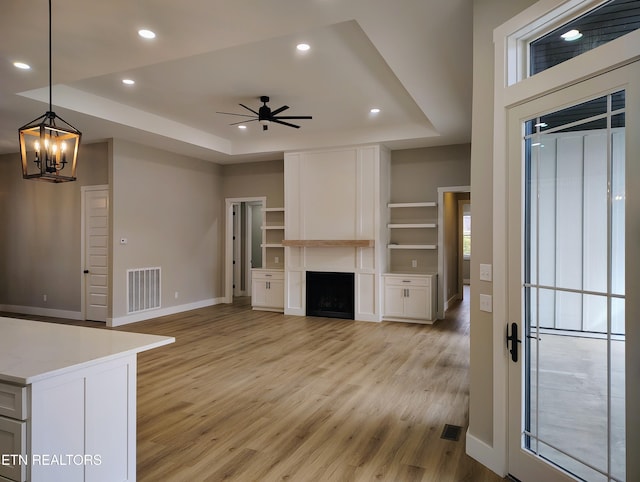 unfurnished living room featuring ceiling fan with notable chandelier, a raised ceiling, light hardwood / wood-style flooring, built in features, and a large fireplace