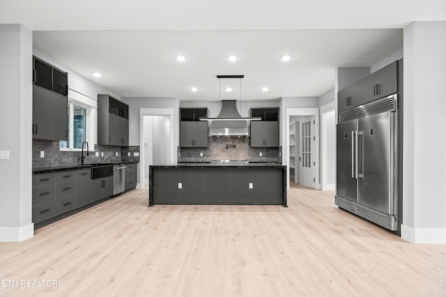 kitchen with pendant lighting, wall chimney range hood, light hardwood / wood-style flooring, a kitchen island, and stainless steel appliances