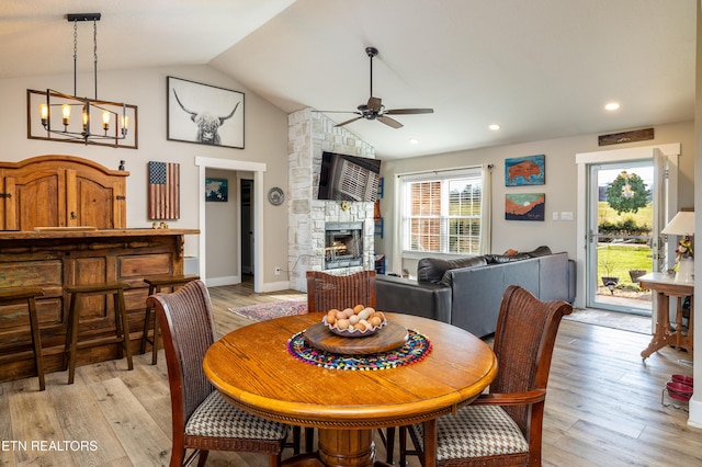 dining area with ceiling fan with notable chandelier, light hardwood / wood-style floors, a stone fireplace, and vaulted ceiling