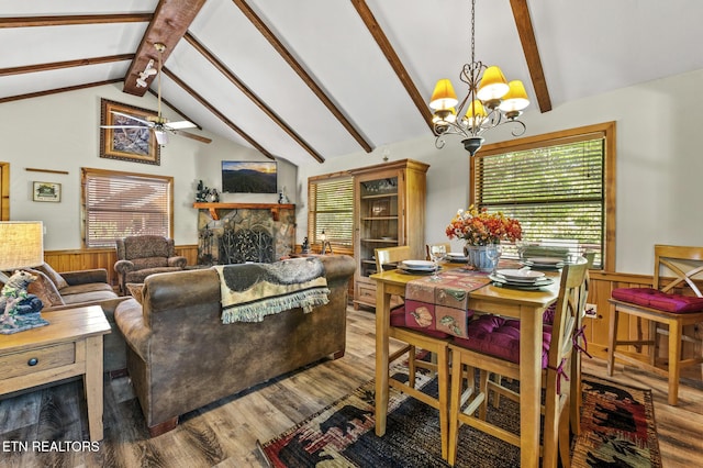 dining room featuring hardwood / wood-style floors, wood walls, ceiling fan with notable chandelier, a stone fireplace, and vaulted ceiling with beams