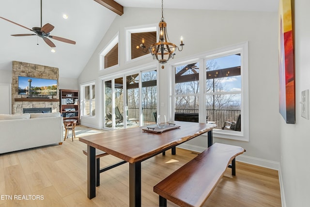 dining area featuring ceiling fan with notable chandelier, light hardwood / wood-style flooring, high vaulted ceiling, beamed ceiling, and a stone fireplace