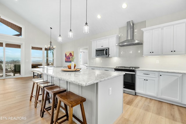 kitchen with white cabinets, a center island with sink, wall chimney range hood, hanging light fixtures, and appliances with stainless steel finishes