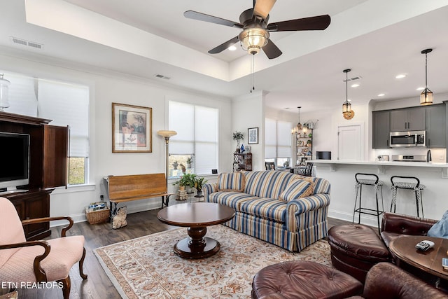 living room featuring ceiling fan with notable chandelier, ornamental molding, dark wood-type flooring, and a tray ceiling