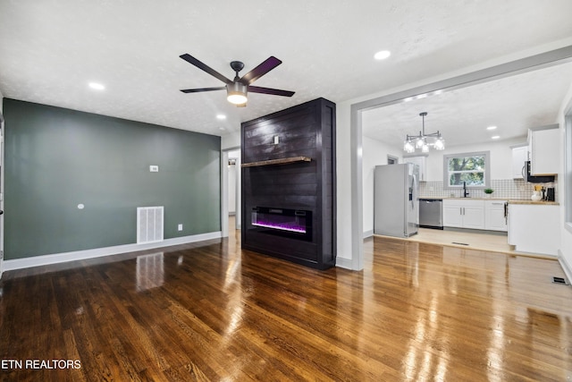 unfurnished living room with sink, a large fireplace, ceiling fan with notable chandelier, and hardwood / wood-style flooring