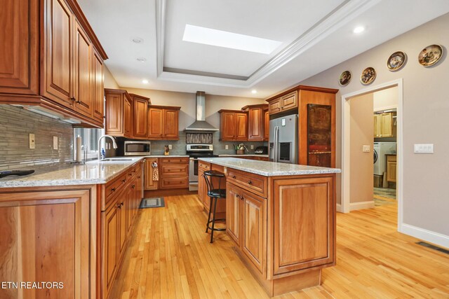 kitchen with a skylight, a center island, wall chimney exhaust hood, a tray ceiling, and appliances with stainless steel finishes