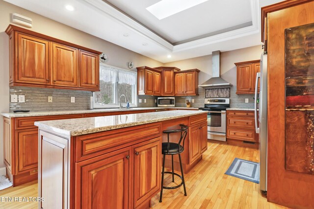 kitchen with a skylight, a center island, wall chimney exhaust hood, a tray ceiling, and appliances with stainless steel finishes