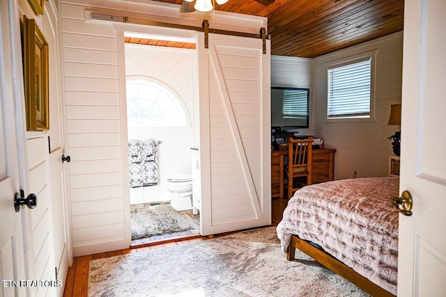 bedroom with wood-type flooring, wood walls, connected bathroom, wooden ceiling, and a barn door