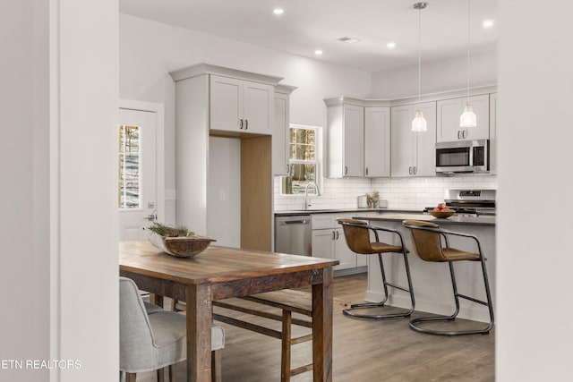 kitchen with stainless steel appliances, wood finished floors, hanging light fixtures, backsplash, and dark countertops