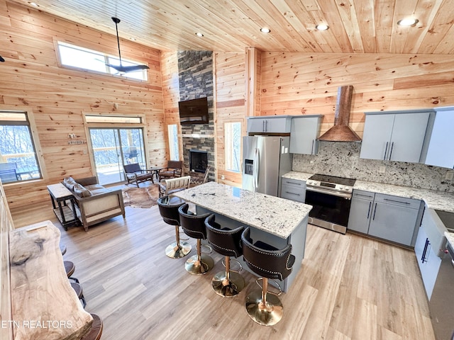 kitchen with wood ceiling, appliances with stainless steel finishes, open floor plan, light stone counters, and wall chimney range hood