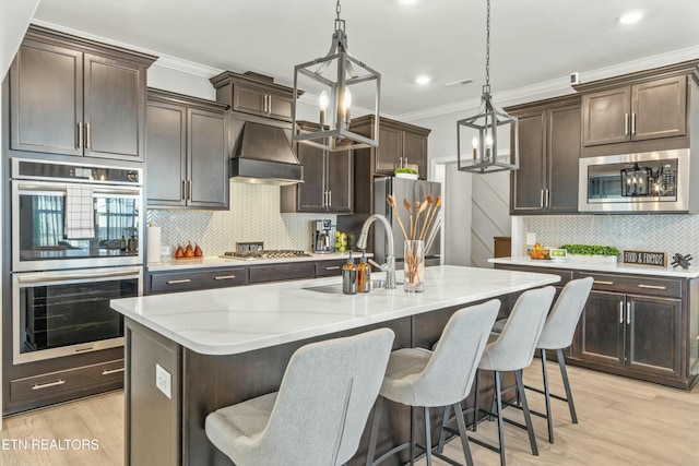 kitchen featuring a kitchen island with sink, sink, appliances with stainless steel finishes, decorative light fixtures, and custom range hood