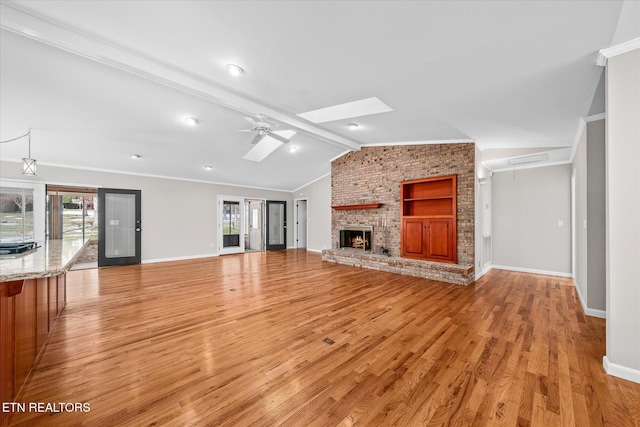 unfurnished living room featuring light wood-type flooring, ceiling fan, a fireplace, vaulted ceiling with skylight, and built in features