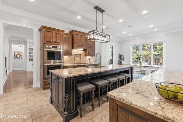 kitchen with stainless steel appliances, ornamental molding, light tile patterned floors, light stone countertops, and a center island with sink