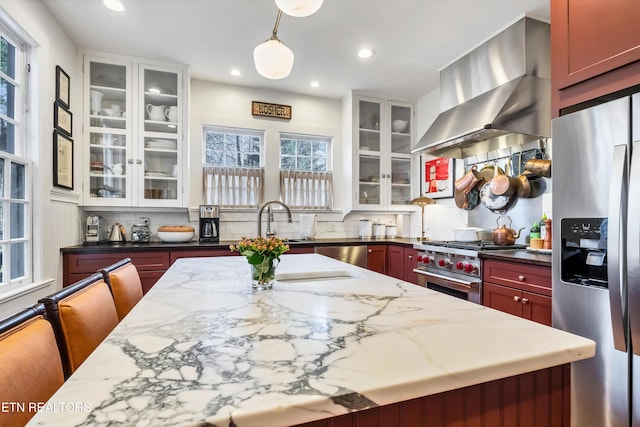 kitchen featuring decorative backsplash, appliances with stainless steel finishes, light stone counters, wall chimney exhaust hood, and pendant lighting