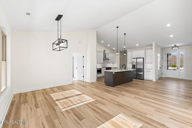 kitchen featuring white cabinetry, wall chimney exhaust hood, stainless steel fridge with ice dispenser, an island with sink, and decorative light fixtures