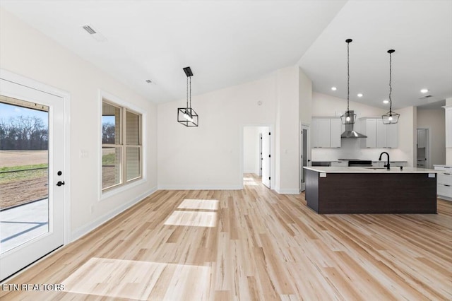 kitchen with light wood-type flooring, wall chimney exhaust hood, vaulted ceiling, pendant lighting, and a center island with sink