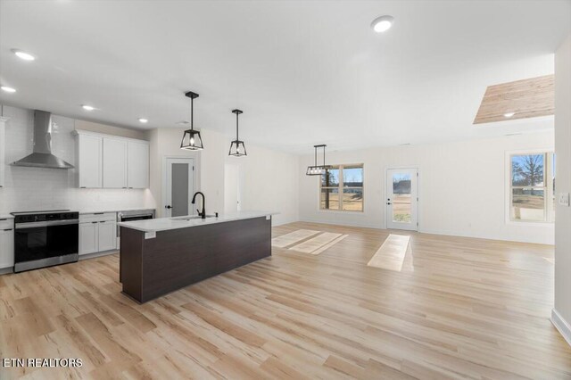 kitchen with light wood-type flooring, wall chimney exhaust hood, white cabinets, stainless steel range with electric cooktop, and an island with sink