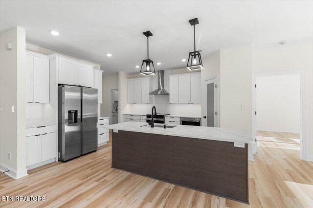 kitchen featuring pendant lighting, wall chimney exhaust hood, stainless steel fridge, an island with sink, and white cabinetry