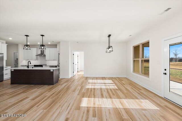 kitchen featuring pendant lighting, a kitchen island with sink, white cabinets, wall chimney range hood, and stainless steel fridge