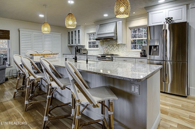 kitchen featuring premium range hood, light stone countertops, appliances with stainless steel finishes, white cabinetry, and a breakfast bar area