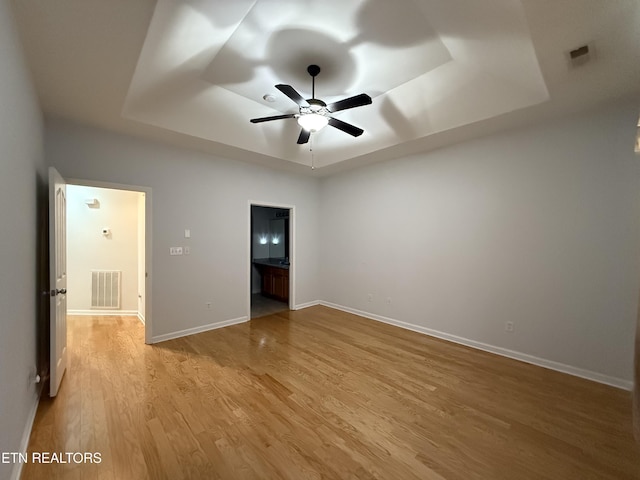 unfurnished bedroom featuring a tray ceiling, ensuite bath, ceiling fan, and light hardwood / wood-style flooring