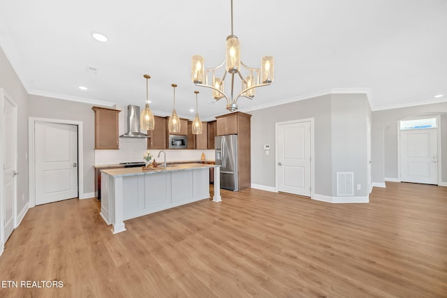 kitchen featuring light stone counters, wall chimney exhaust hood, stainless steel appliances, a kitchen island with sink, and pendant lighting