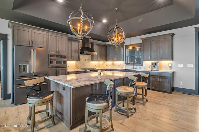 kitchen featuring stainless steel appliances, a tray ceiling, wall chimney range hood, decorative light fixtures, and a large island