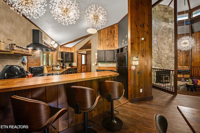 kitchen featuring wall chimney range hood, a notable chandelier, butcher block countertops, a breakfast bar, and black appliances