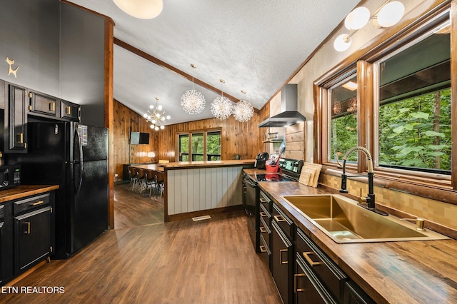 kitchen featuring wood counters, black appliances, wall chimney range hood, lofted ceiling with beams, and hanging light fixtures