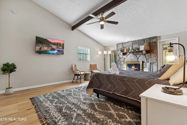 bedroom featuring a textured ceiling, ceiling fan, wood-type flooring, lofted ceiling with beams, and a stone fireplace