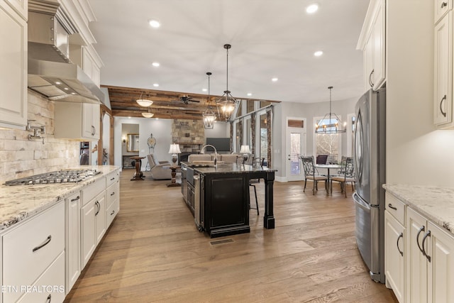 kitchen with light stone countertops, appliances with stainless steel finishes, wall chimney exhaust hood, hanging light fixtures, and a kitchen island with sink