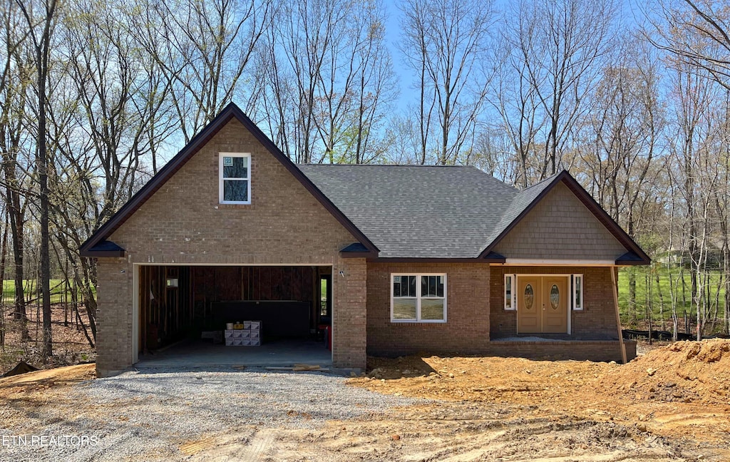 view of front of house with gravel driveway, roof with shingles, covered porch, brick siding, and a garage
