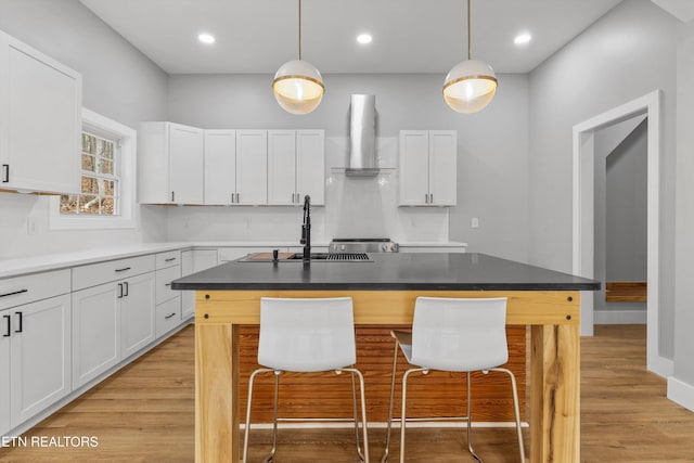 kitchen with wall chimney range hood, sink, an island with sink, and white cabinets