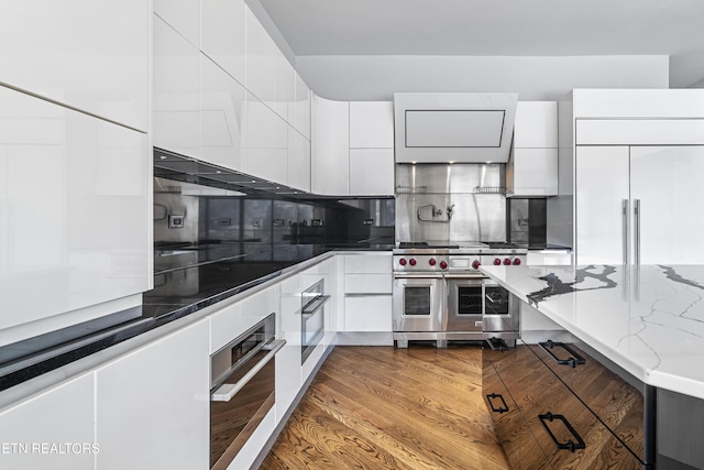 kitchen with tasteful backsplash, high end appliances, dark stone countertops, white cabinets, and hardwood / wood-style floors