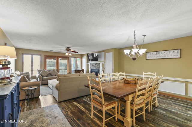 dining space featuring dark hardwood / wood-style flooring, a stone fireplace, ceiling fan with notable chandelier, and a textured ceiling