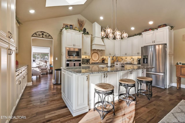 kitchen with custom exhaust hood, backsplash, a center island with sink, hanging light fixtures, and appliances with stainless steel finishes