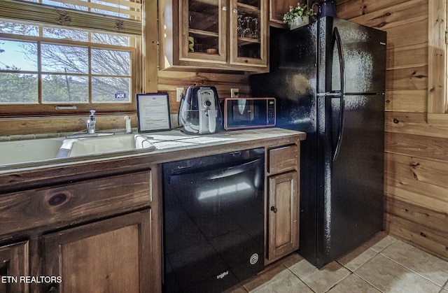 kitchen with sink, dishwasher, black refrigerator, light tile patterned floors, and wood walls