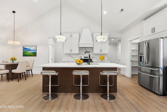 kitchen featuring white cabinetry, custom range hood, an island with sink, hanging light fixtures, and appliances with stainless steel finishes