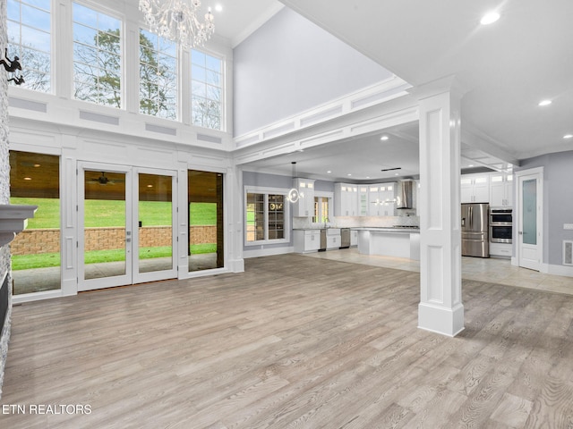 unfurnished living room featuring a towering ceiling, ornamental molding, light hardwood / wood-style flooring, an inviting chandelier, and decorative columns