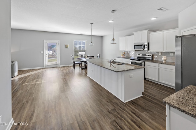 kitchen with appliances with stainless steel finishes, a center island with sink, pendant lighting, white cabinetry, and dark stone countertops