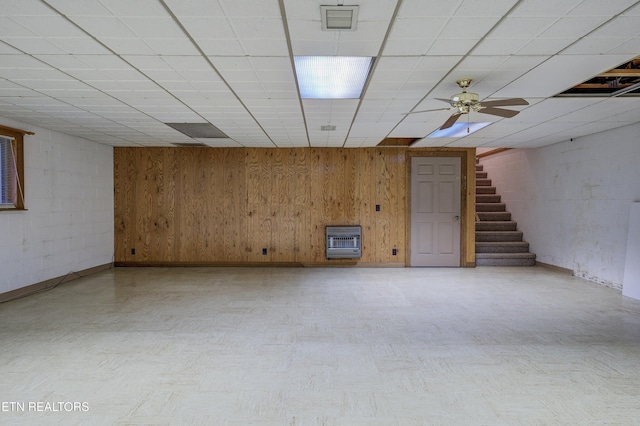 basement with ceiling fan, wooden walls, heating unit, and a drop ceiling