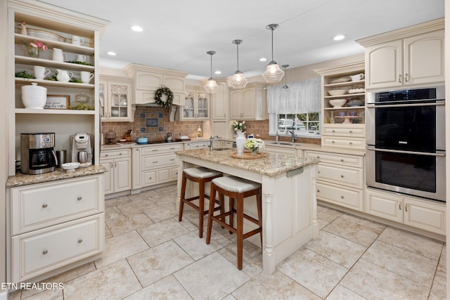 kitchen featuring cream cabinetry, decorative light fixtures, light stone countertops, a kitchen island, and double oven