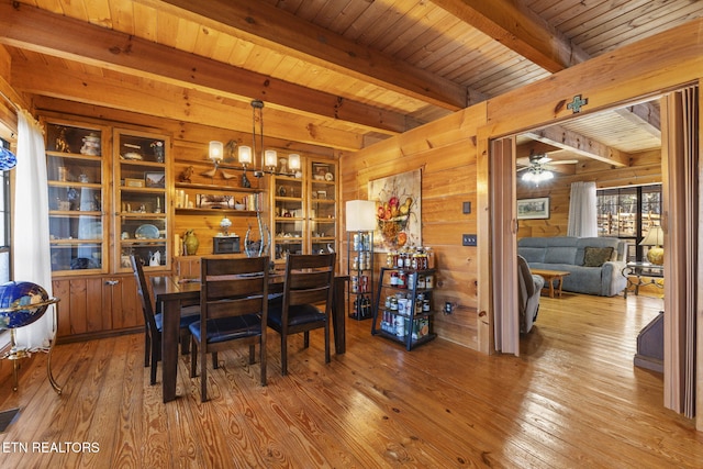 dining space with ceiling fan with notable chandelier, hardwood / wood-style floors, wooden walls, and beamed ceiling