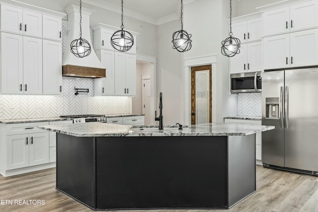 kitchen featuring appliances with stainless steel finishes, white cabinetry, premium range hood, hanging light fixtures, and a kitchen island with sink